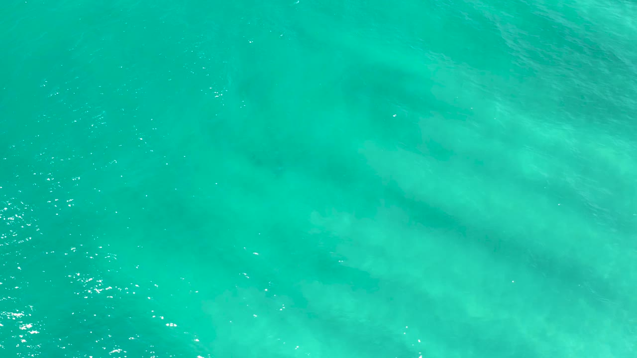 Aerial frame of blue-green water in San Jose Del Cabo with soft pale subsurface shapes spreading across the center and no visible people.