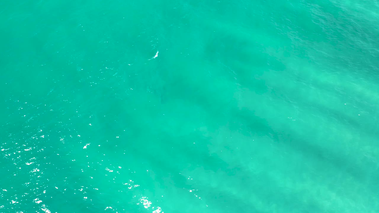 Top-down turquoise sea in San Jose Del Cabo with pale underwater streaks and brighter sun sparkles along the right side, with no visible people.