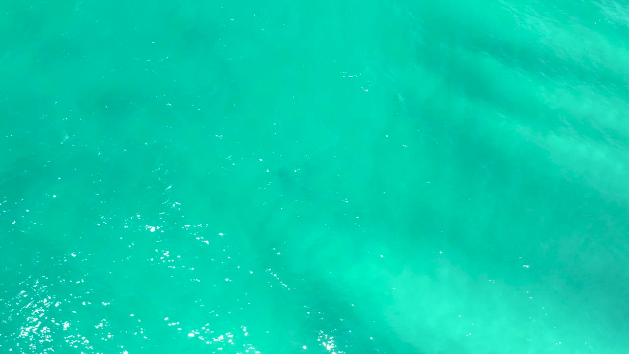 Bright turquoise sea surface in San Jose del Cabo shows scattered white sun reflections and soft cloudy shapes below the water, with no visible people.