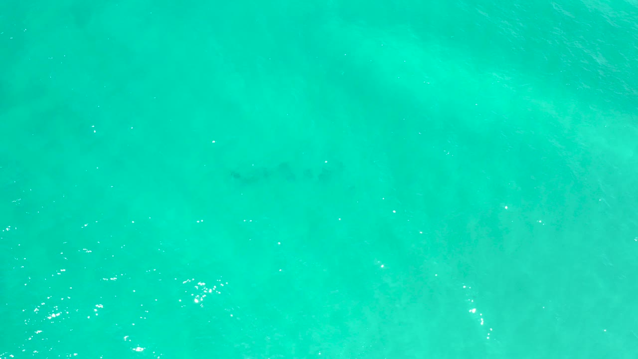 Overhead view of turquoise water in San Jose Del Cabo with pale sand-like patches below the surface, scattered sun glints, and no visible people.