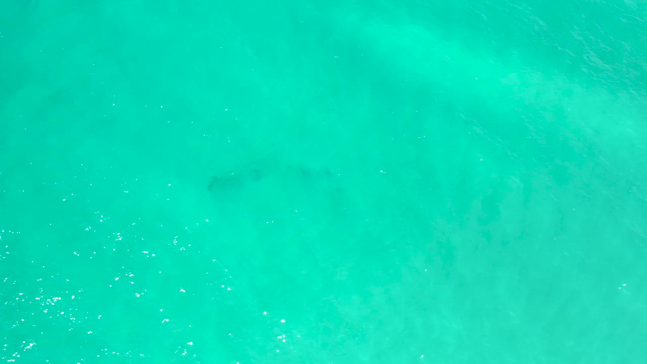 Bright aqua sea surface in San Jose Del Cabo shows uneven lighter and darker underwater areas, tiny sun flecks, and no visible people.