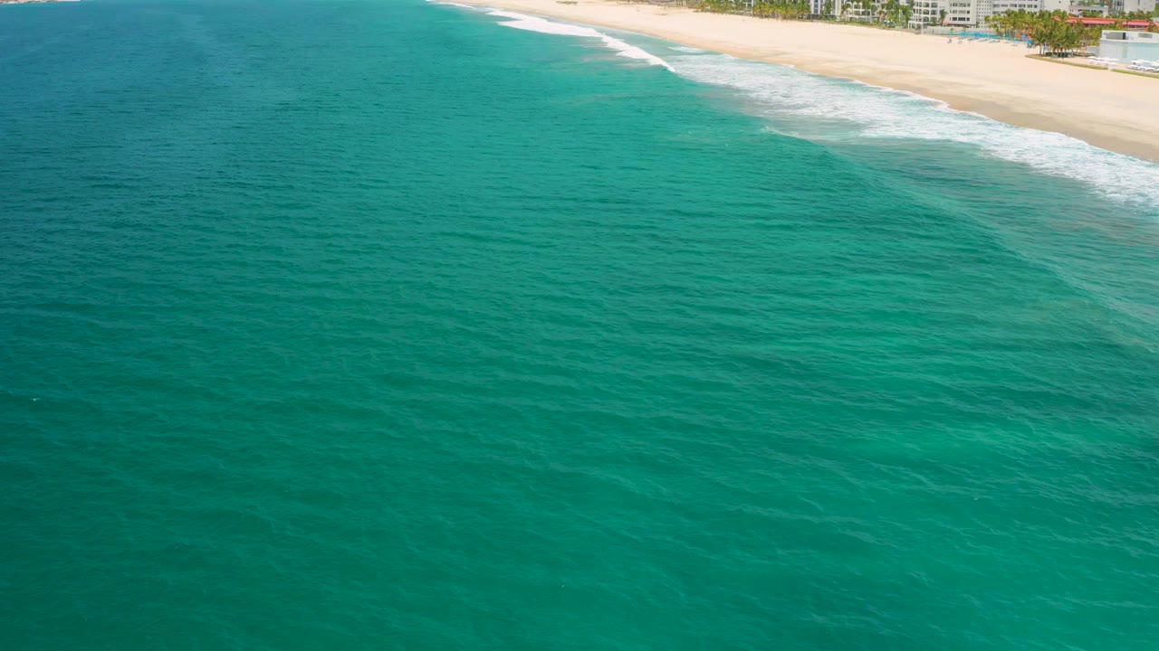 Aerial view in San Jose Del Cabo with turquoise water filling the foreground, pale beach and white surf at the top edge, and no visible people.