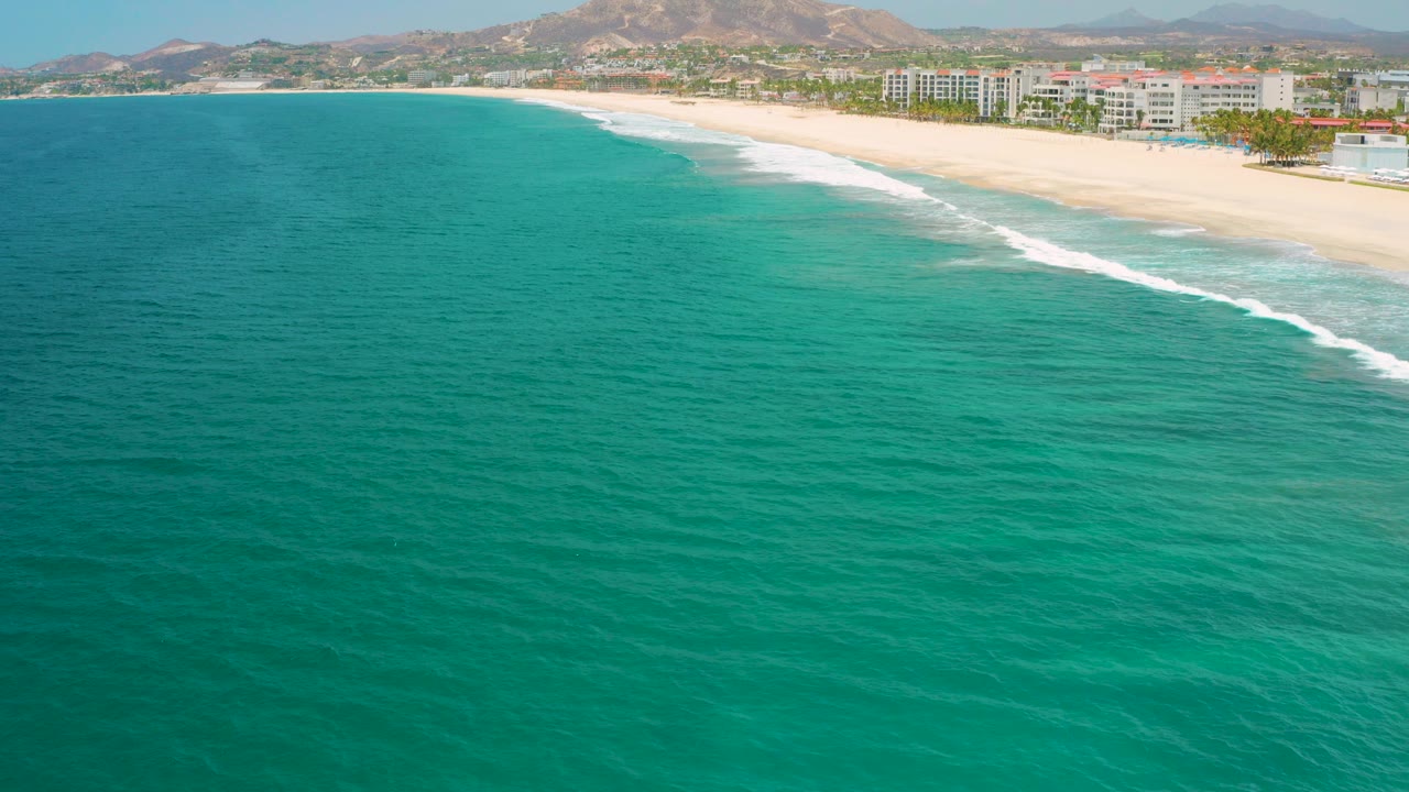 Opening aerial view in San Jose Del Cabo, Mexico with turquoise water in the foreground, a pale beach curving right, resort buildings behind it, dry hills in the distance, and no visible people.
