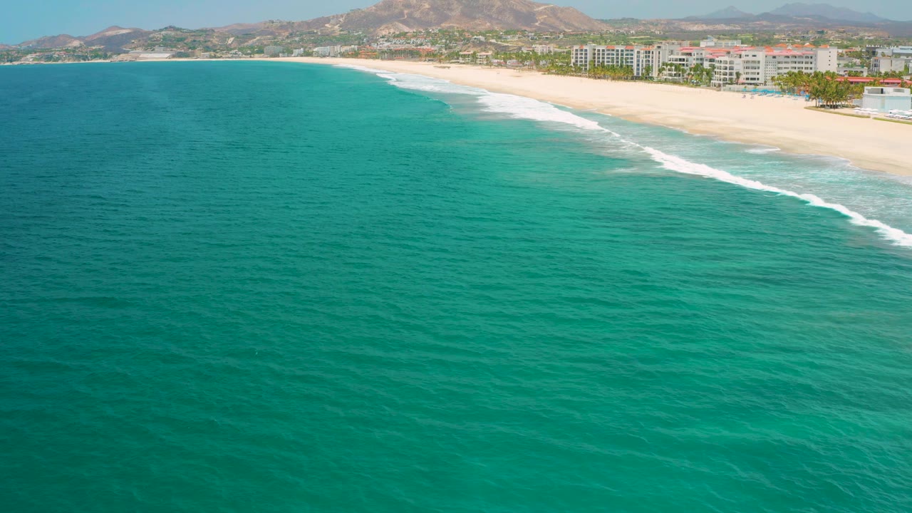 A wide San Jose Del Cabo beach frame shows more open turquoise water across the lower frame, white surf tracing the sand, resort buildings near the right shoreline, distant hills, and no visible people.