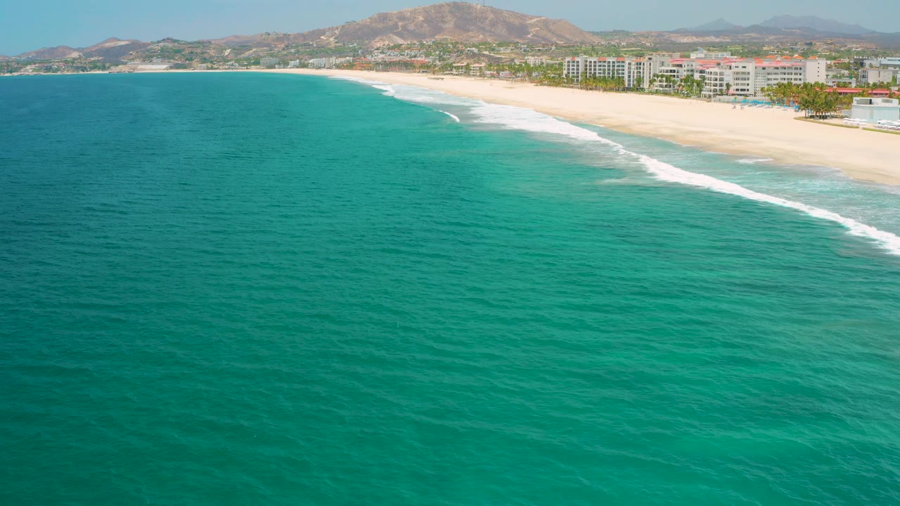 The San Jose Del Cabo shoreline curves across the upper frame, with textured turquoise ocean below, a white wave band along the beach, resort blocks set back from the sand, hills beyond, and no visible people.