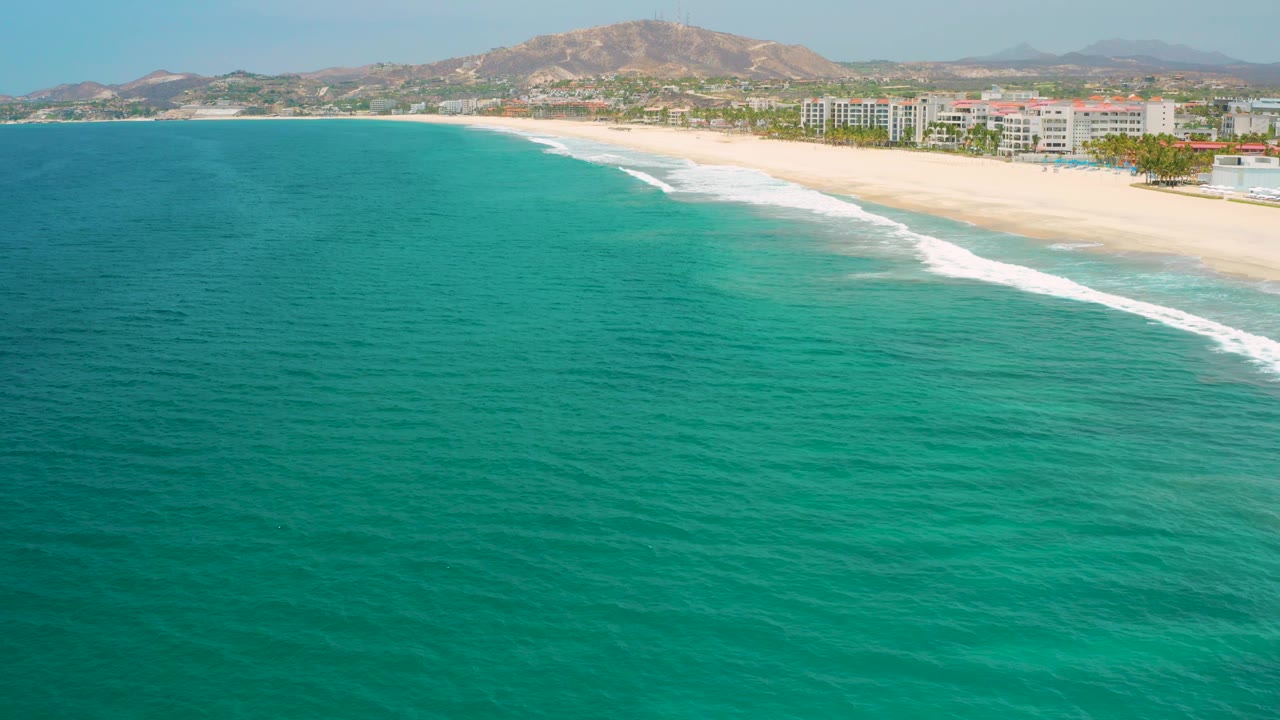 Diagonal aerial view of beach surf with turquoise water and resort frontage.