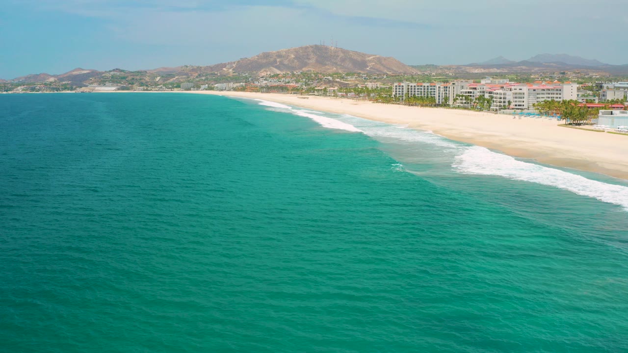 Opening aerial frame over turquoise water and a pale beach in San Jose Del Cabo, Mexico, with resort buildings and low hills beyond the shore, no visible people.