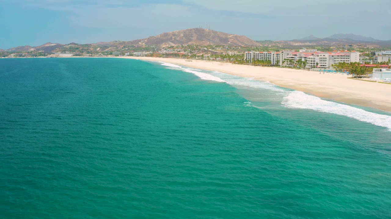 Aerial frame one second in shows San Jose Del Cabo shoreline with green-blue ocean dominating the foreground, a diagonal surf line, resort buildings, hills, and no visible people.