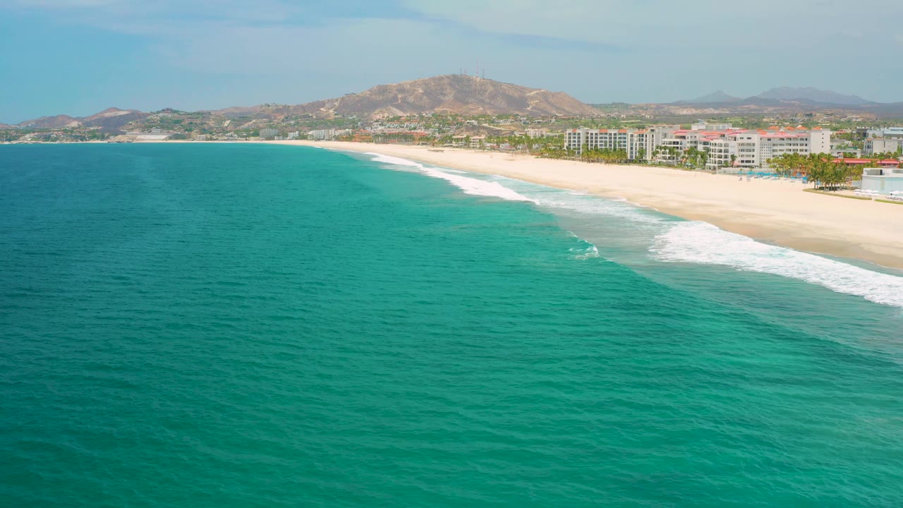 Two seconds in, the aerial view holds on the curving San Jose Del Cabo beach as white waves trace the sand below hotel buildings, with no visible people.