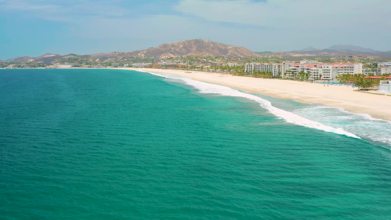 San Jose Del Cabo aerial frame with turquoise water covering most of the foreground, a long breaking wave near shore, white beachfront buildings to the right, hills behind, and no visible people.