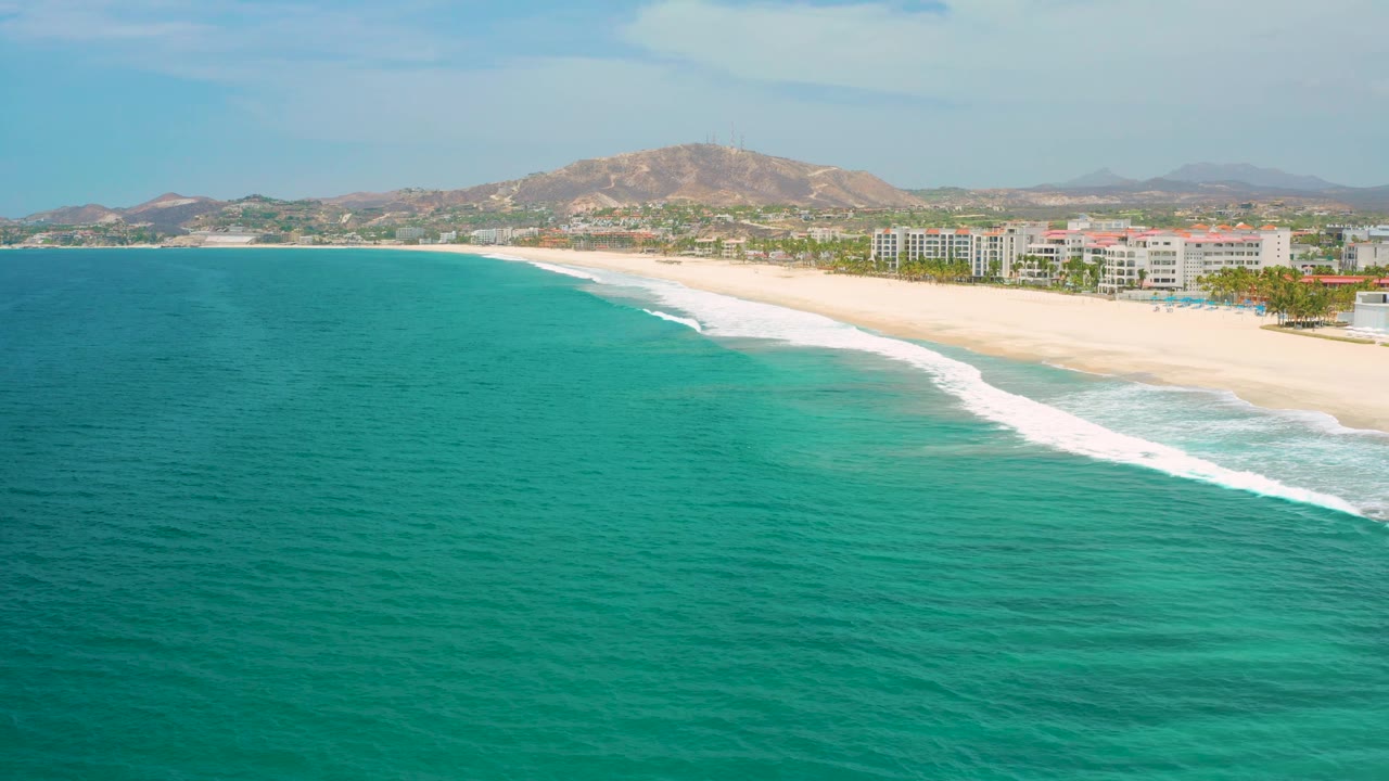 San Jose Del Cabo coastline frame with a broad blue-green ocean foreground, white waves curling along the beach, compact shoreline buildings on the right, hazy hills behind them, and no visible people.