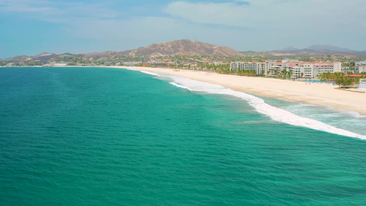 Mid-clip frame of San Jose Del Cabo beach with textured blue water in front, foam bands reaching the sand, white buildings along the shore, brown hills in back, and no visible people.