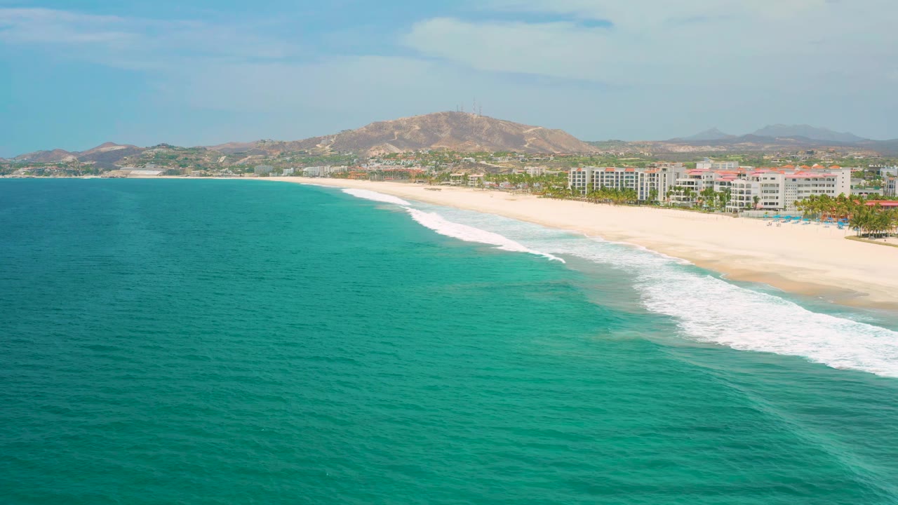 Wide aerial frame in San Jose Del Cabo, Mexico, showing a longer sweep of turquoise water, white wave lines meeting the sand, shorefront buildings, dry hills, light cloud cover, and no visible people.