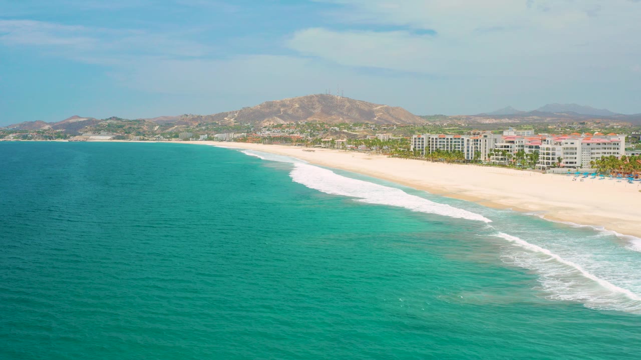 Wide opening aerial frame over turquoise water toward a sandy San Jose del Cabo beach, with white surf, shoreline buildings, palms, tan hills, and no visible people.