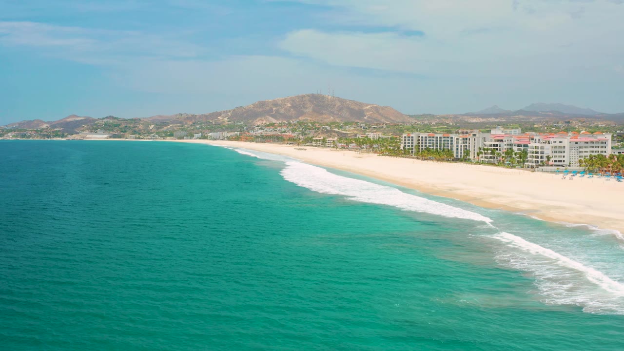 Aerial view along the San Jose del Cabo coast with more open turquoise water in the foreground, pale sand at right, low buildings beyond the beach, hills, and no visible people.