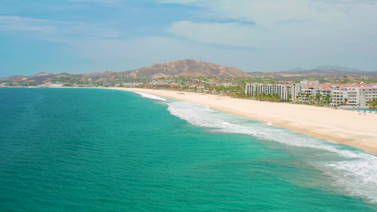 Aerial San Jose Del Cabo coastline frame with the surf line running diagonally across pale sand, turquoise sea covering the left foreground, low buildings on the right, hills behind, and no visible people.