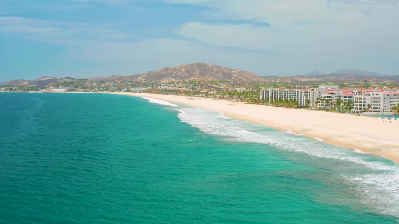 Aerial frame over San Jose Del Cabo, Mexico showing more open turquoise water in front, a foamy surf band at the beach edge, resort-like shoreline buildings, tan hills, and no visible people.