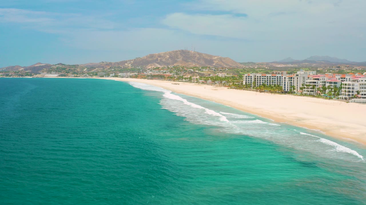 Aerial frame over San Jose Del Cabo showing turquoise water in the foreground, a pale beach with breaking surf, resort buildings, low hills, and no visible people.