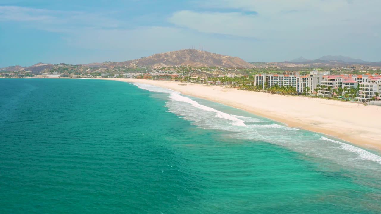 San Jose Del Cabo shoreline from above, with darker teal water at left, white waves rolling onto the pale beach, resort blocks near the right edge, inland hills, and no visible people.