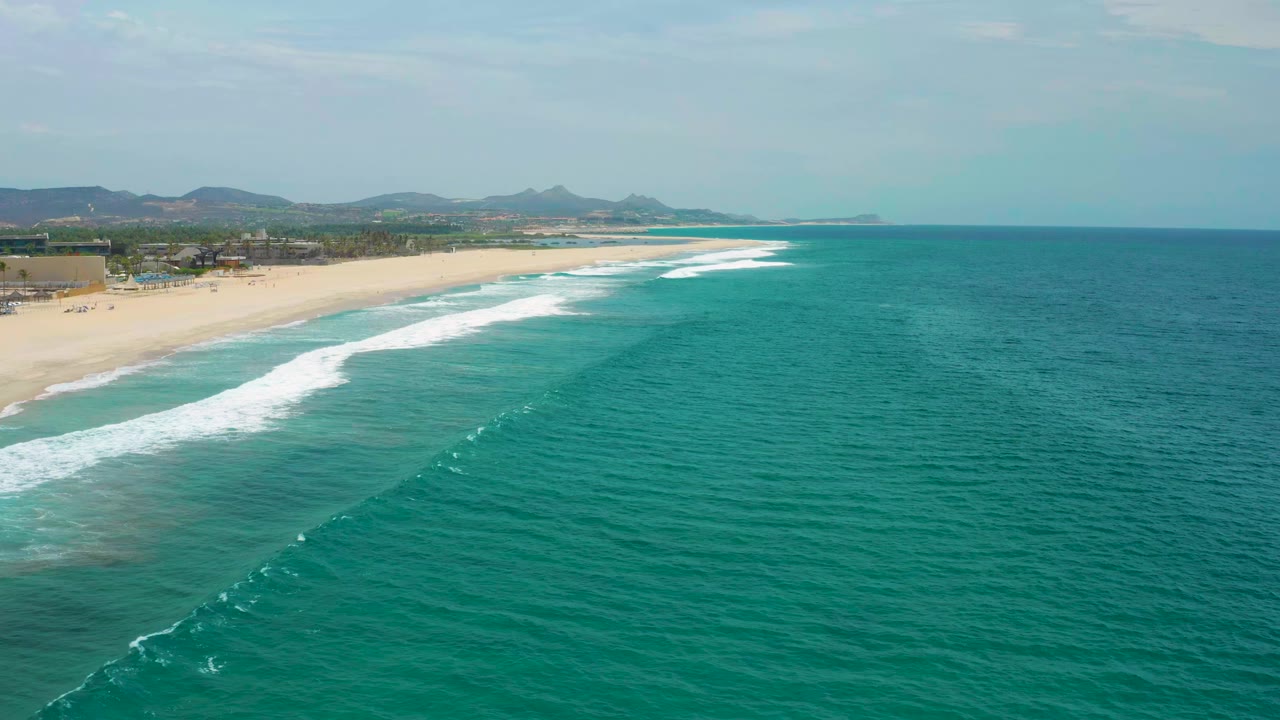 San Jose Del Cabo coastline with a diagonal breaking wave across the turquoise ocean, golden sand along the left edge, low buildings inland, and no visible people.