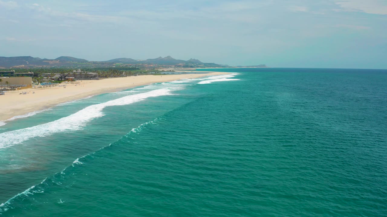 Wide aerial view of San Jose Del Cabo, Mexico, with open blue-green water dominating the frame, surf bands near the beach, distant hills, and no visible people.