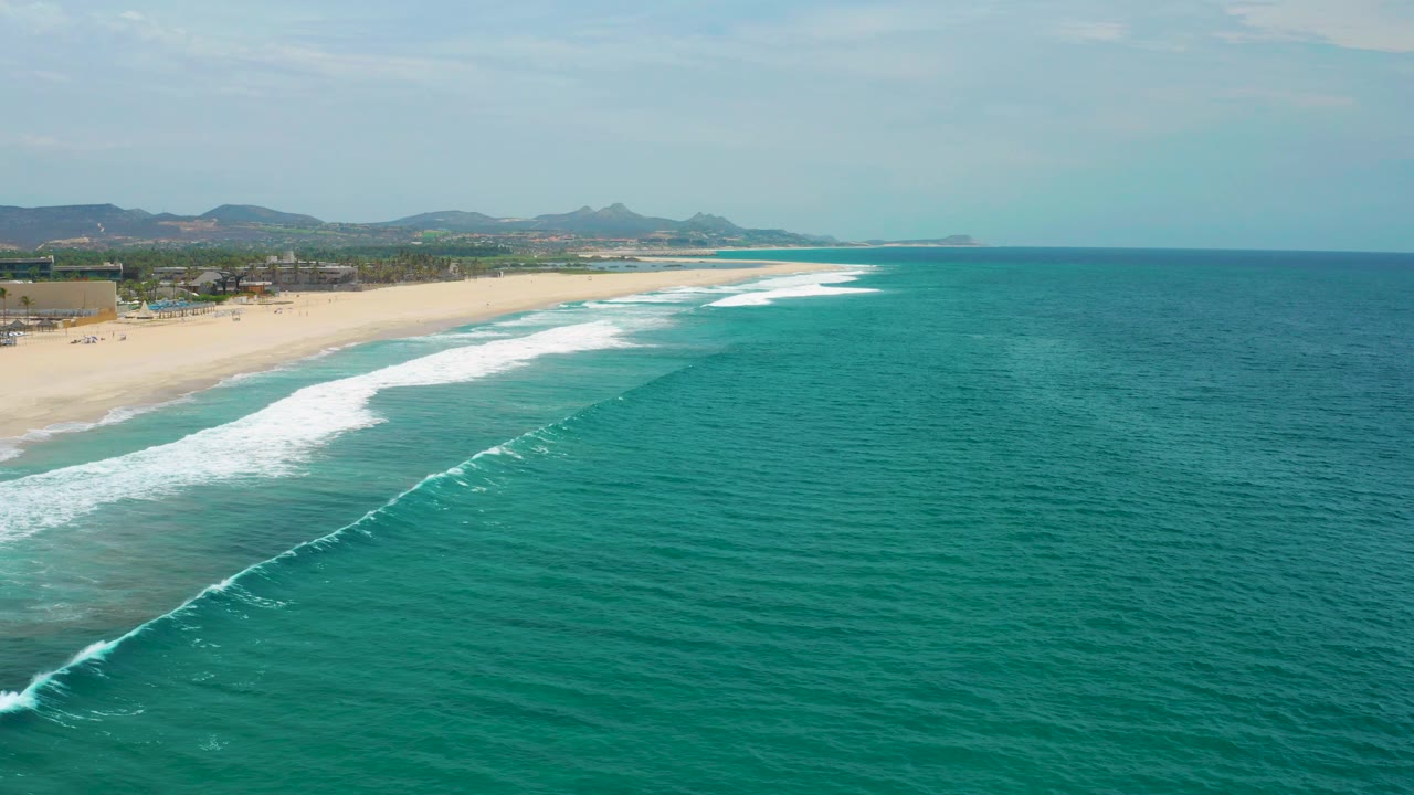 Aerial frame over San Jose Del Cabo showing white waves rolling toward the sandy beach, shallow green water near shore, inland buildings, hills, and no visible people.