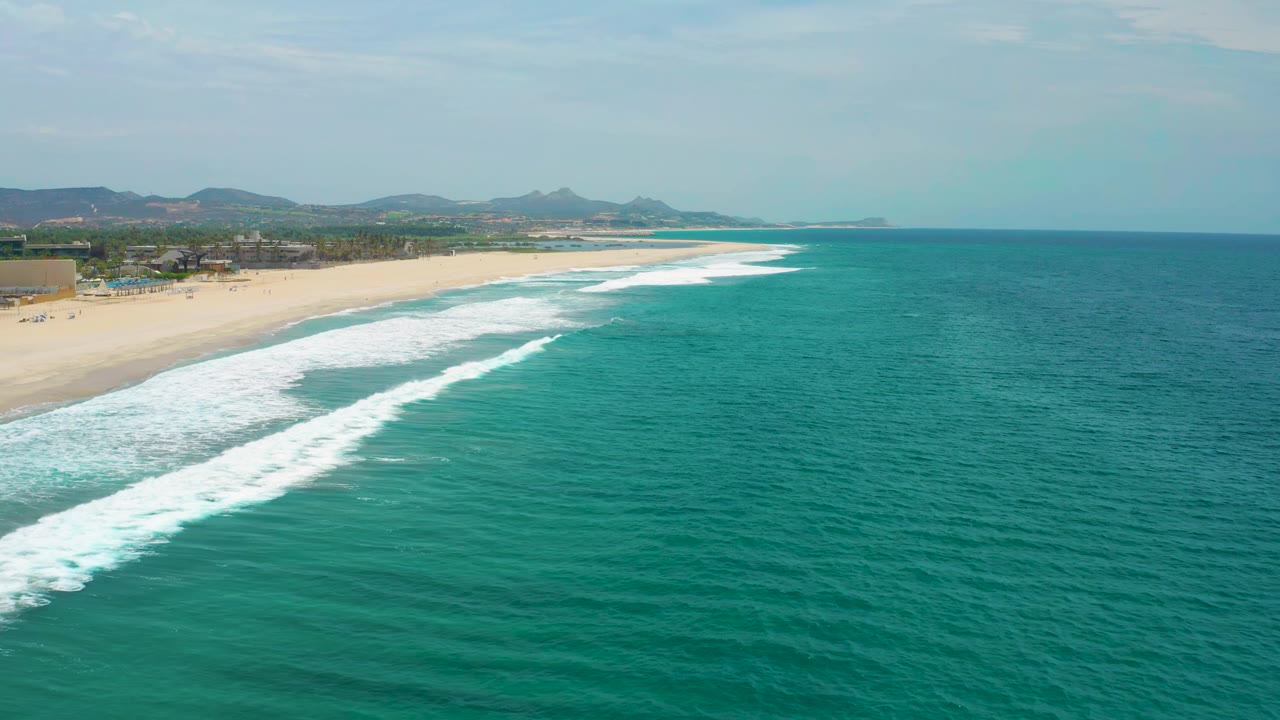 Aerial frame over San Jose del Cabo with long white surf bands advancing toward the beach, turquoise ocean to the right, low hills beyond, and no visible people.