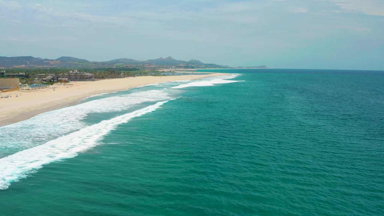 Oblique aerial view of the San Jose del Cabo coast with foamy surf curving along the beach, open blue-green water, distant mountains, and no visible people.