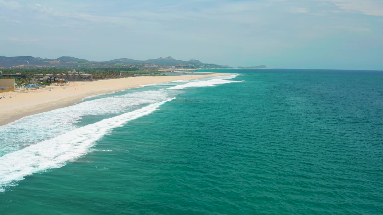 Aerial view of San Jose del Cabo where a broad white wave reaches the lower-left shoreline, with teal water across the frame, distant hills, and no visible people.