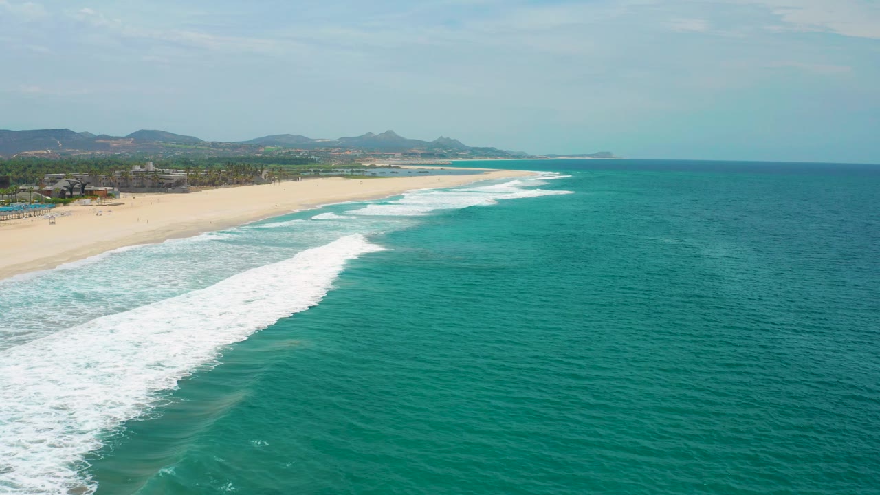 Aerial coastline view in San Jose Del Cabo, Mexico with a long sandy beach, white surf bands, clear green water, distant hills, and no visible people.