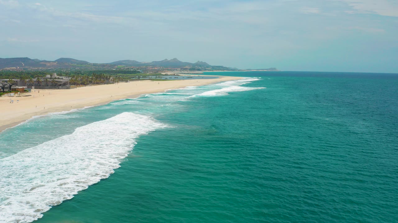 Aerial frame from San Jose Del Cabo, Mexico showing turquoise ocean, white surf angled toward a broad sandy beach, distant hills, and no visible people.
