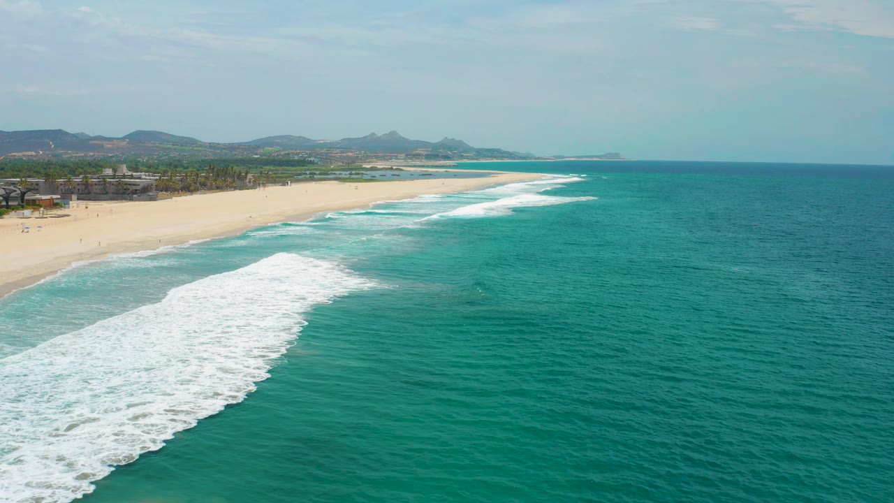 Aerial view in San Jose Del Cabo, Mexico with a foamy wave band closer to the lower left, open teal water to the right, distant hills, and no visible people.
