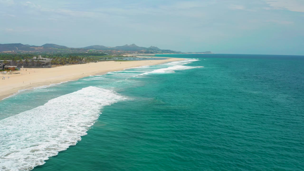 San Jose Del Cabo, Mexico coastline seen from above, with repeated white surf bands along the sand, open turquoise water on the right, hills beyond, and no visible people.