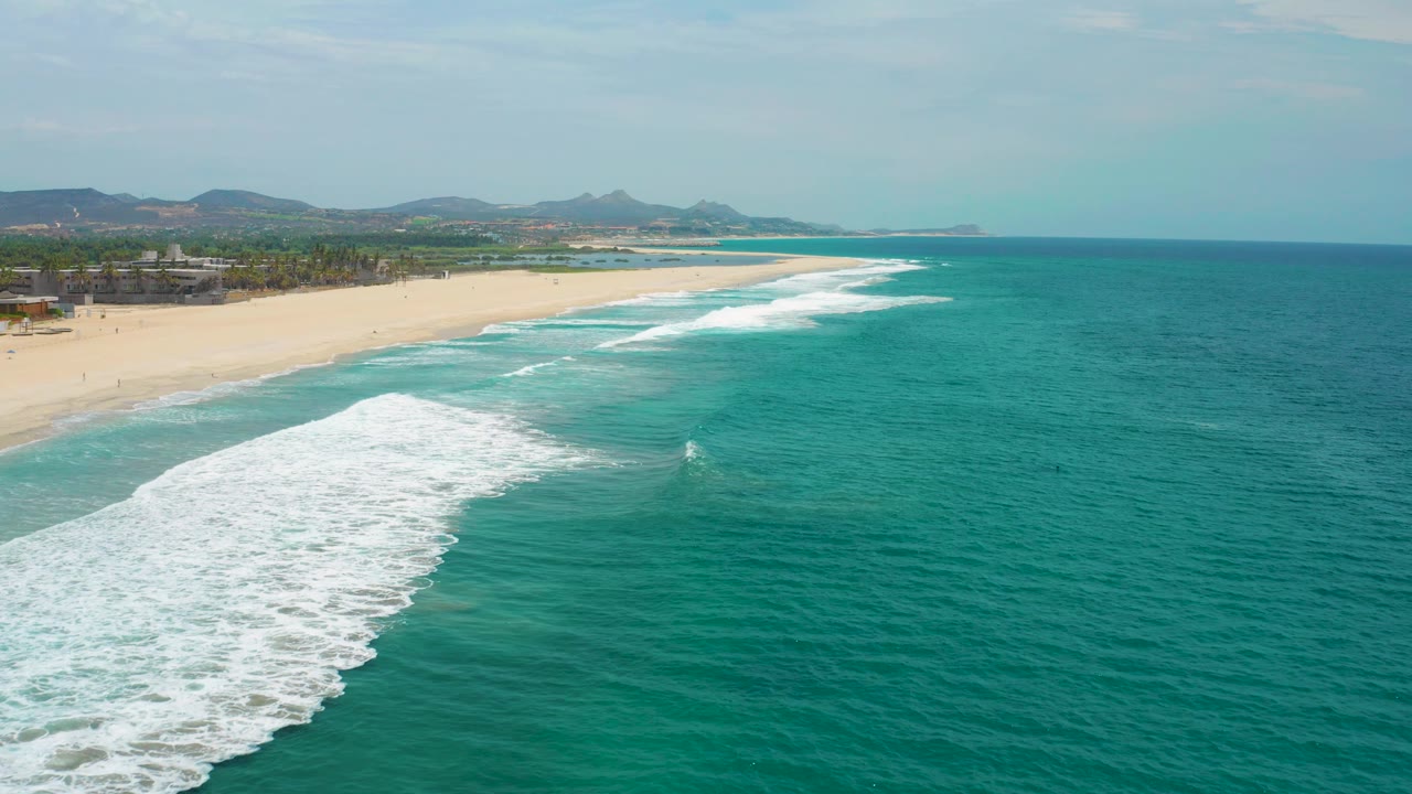 Elevated San Jose Del Cabo, Mexico beach view with a broad white foam patch at lower left, pale sand along the left side, teal ocean to the right, and no visible people.