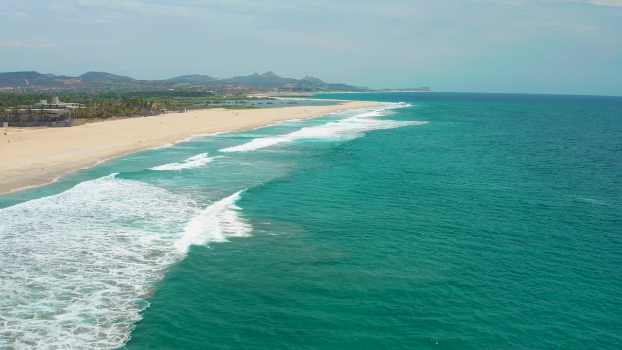 Aerial view in San Jose del Cabo showing turquoise water at right, white surf cutting diagonally toward a long sandy beach at left, distant hills on the horizon, and no visible people.