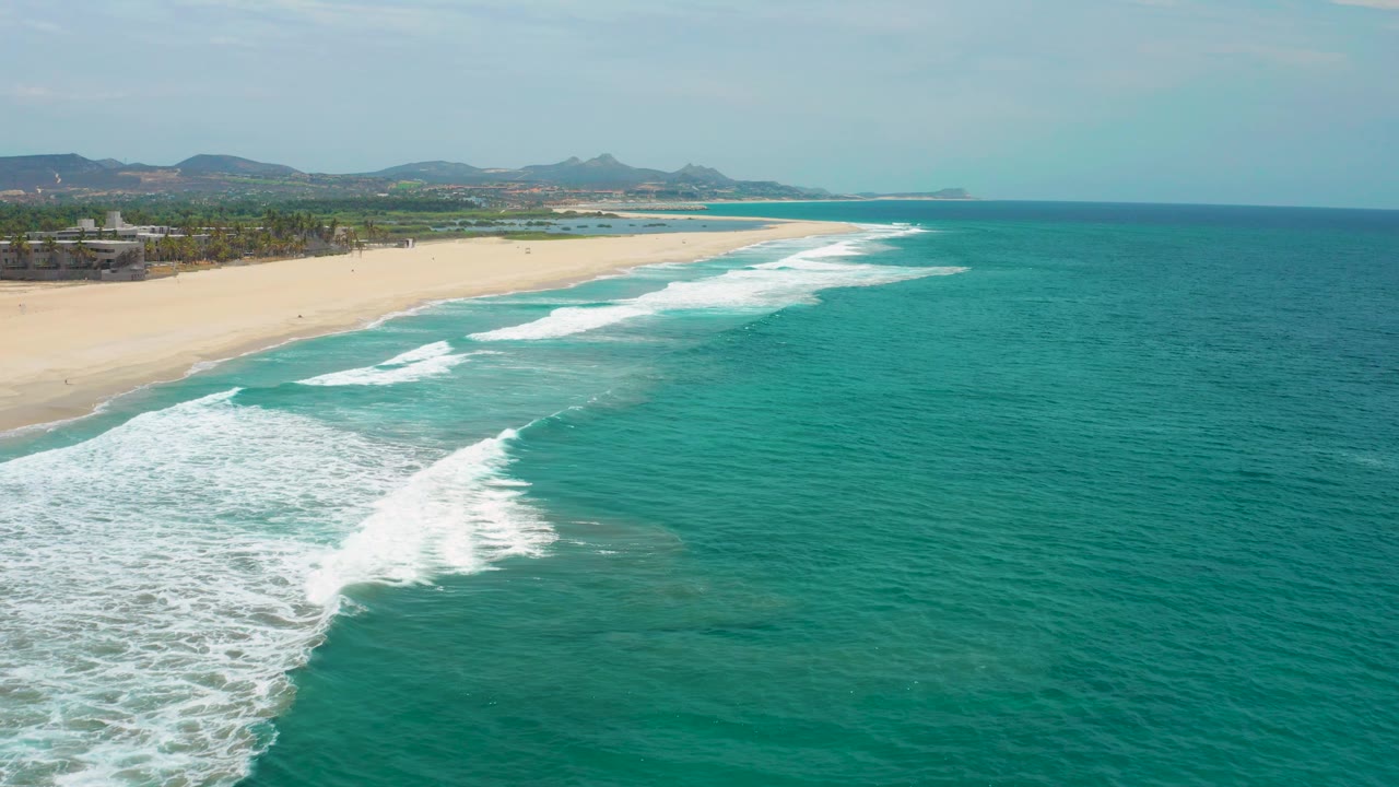 At two seconds, the wave break sits farther along the beach, with textured blue-green ocean filling most of the right side and the San Jose del Cabo coast receding into distant hills, no visible people.