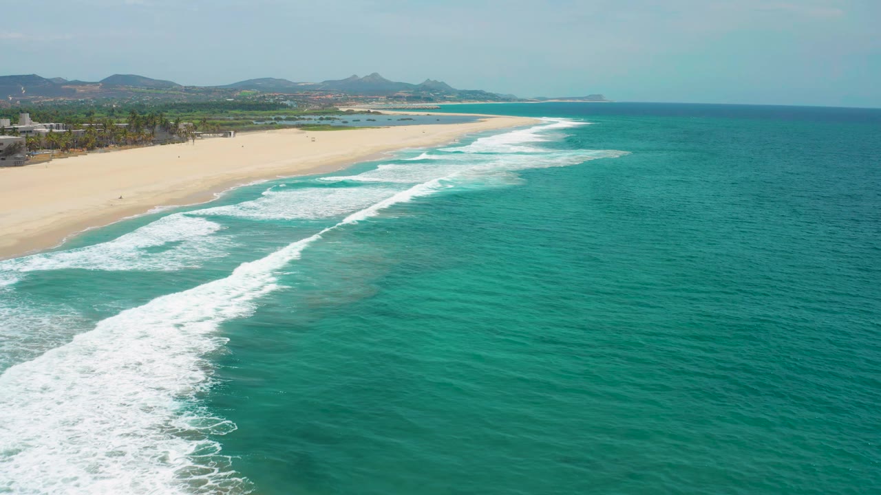 San Jose del Cabo coastline seen from above, with a wider band of white surf crossing the lower left water, the beach running along the upper left, and no visible people.