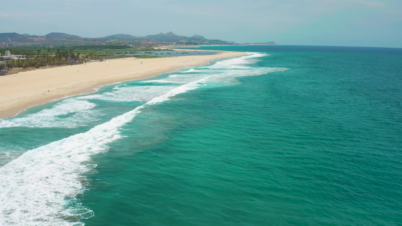 Aerial frame over San Jose del Cabo showing open teal water on the right, foamy wave lines near the left edge, a pale sandy beach, distant hills, and no visible people.