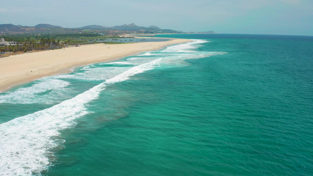 Bright aerial view in San Jose del Cabo with white surf curling across turquoise water, a long sandy beach at left, palm-lined land beyond, and no visible people.