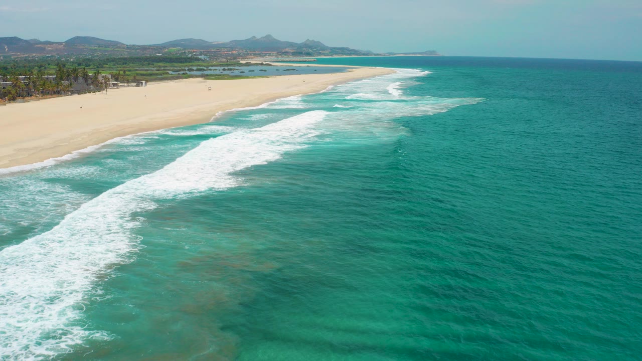Aerial view of San Jose del Cabo, Mexico coastline with turquoise water, white surf reaching a broad empty beach, green land behind it, and no visible people.