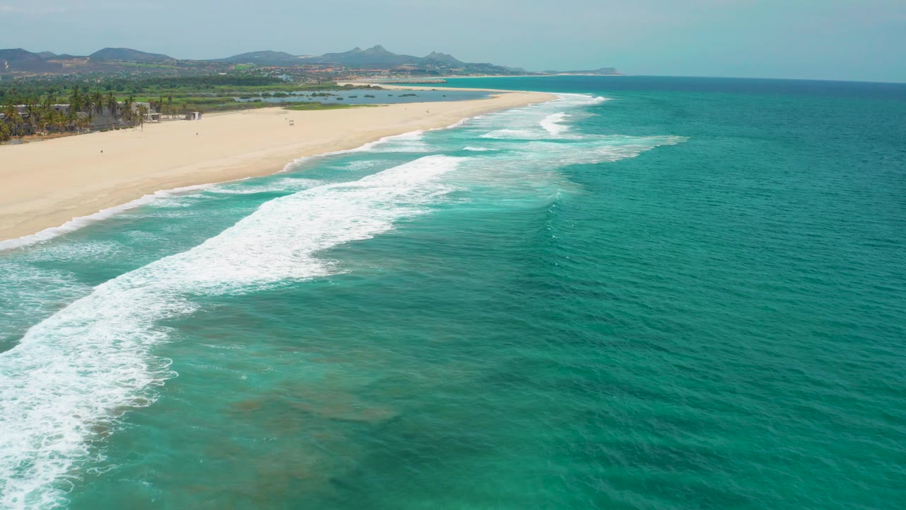Wide aerial frame of San Jose del Cabo beach with a long white wave line, open turquoise water to the right, empty sand to the left, and no visible people.