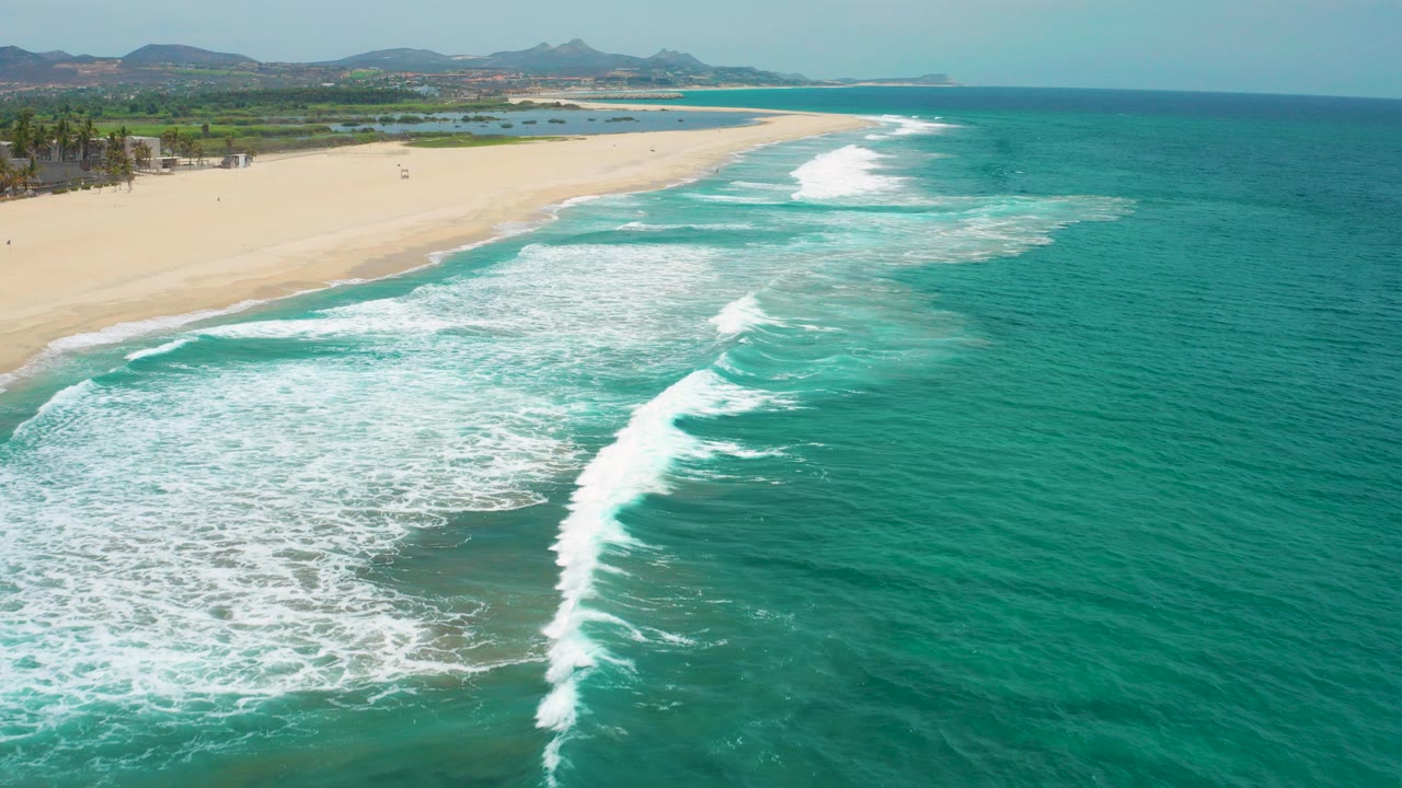 Aerial view over San Jose Del Cabo surf, with turquoise water and white foam approaching a broad sandy beach, no visible people.