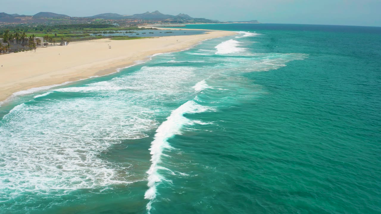 White surf breaks diagonally beside the San Jose Del Cabo beach, with green inland terrain and low hills beyond the sand, no visible people.