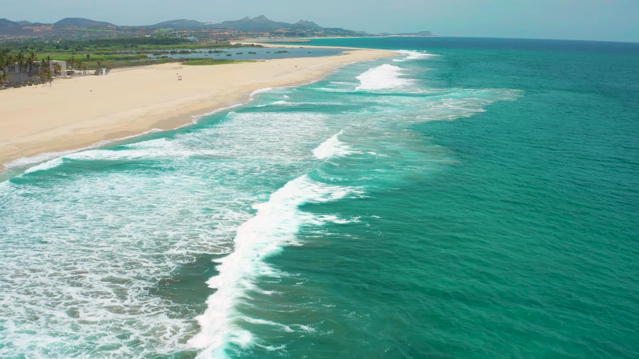 Turquoise water fills the right side while layered foam bands wash toward the pale San Jose Del Cabo shoreline, no visible people.