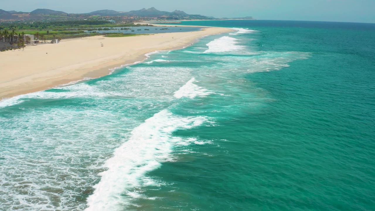 Foamy surf spreads across shallow turquoise water near the San Jose Del Cabo beach, with open sand and low greenery in the background, no visible people.