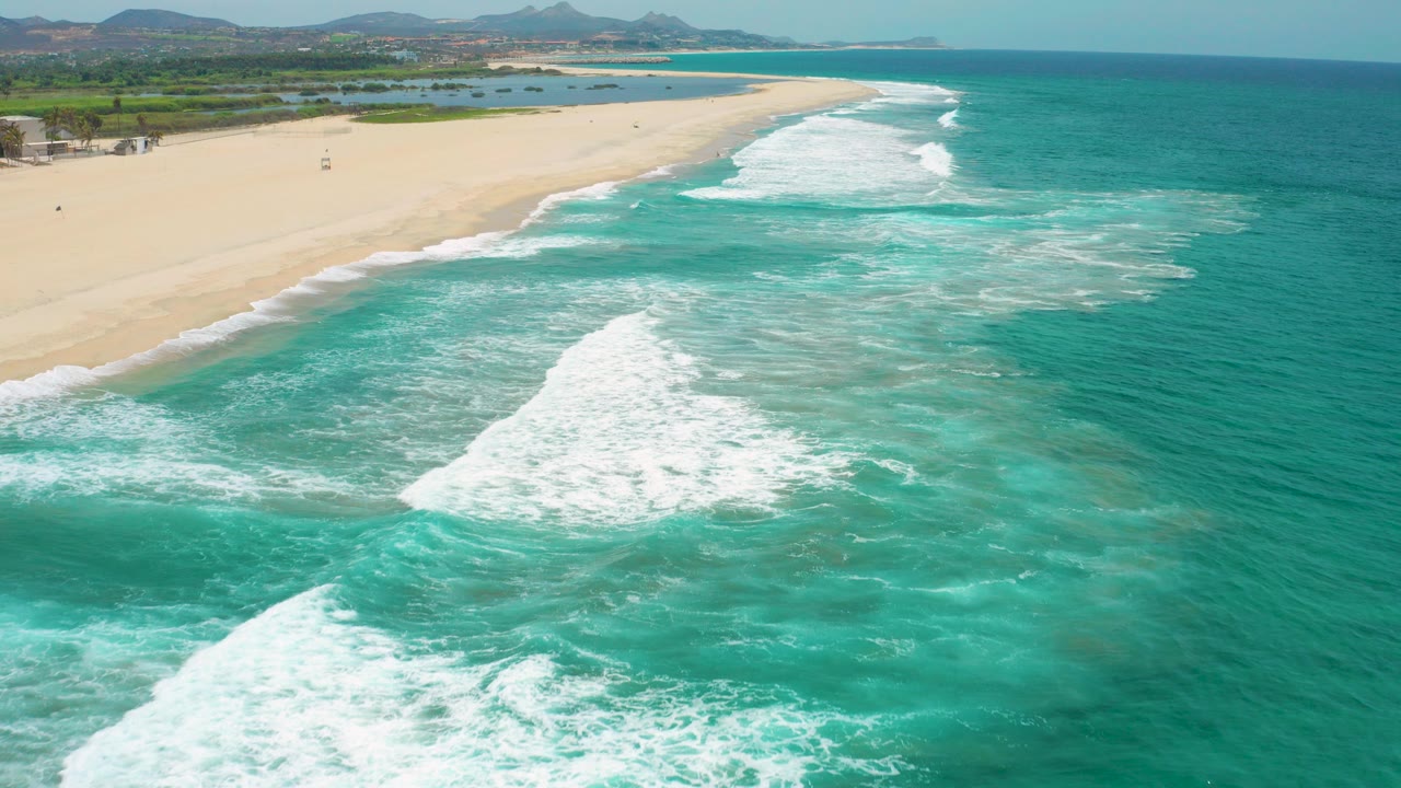 Aerial view over San Jose Del Cabo, Mexico with turquoise water in the foreground, white surf breaking toward a wide sandy beach, distant hills on the horizon, and no visible people.