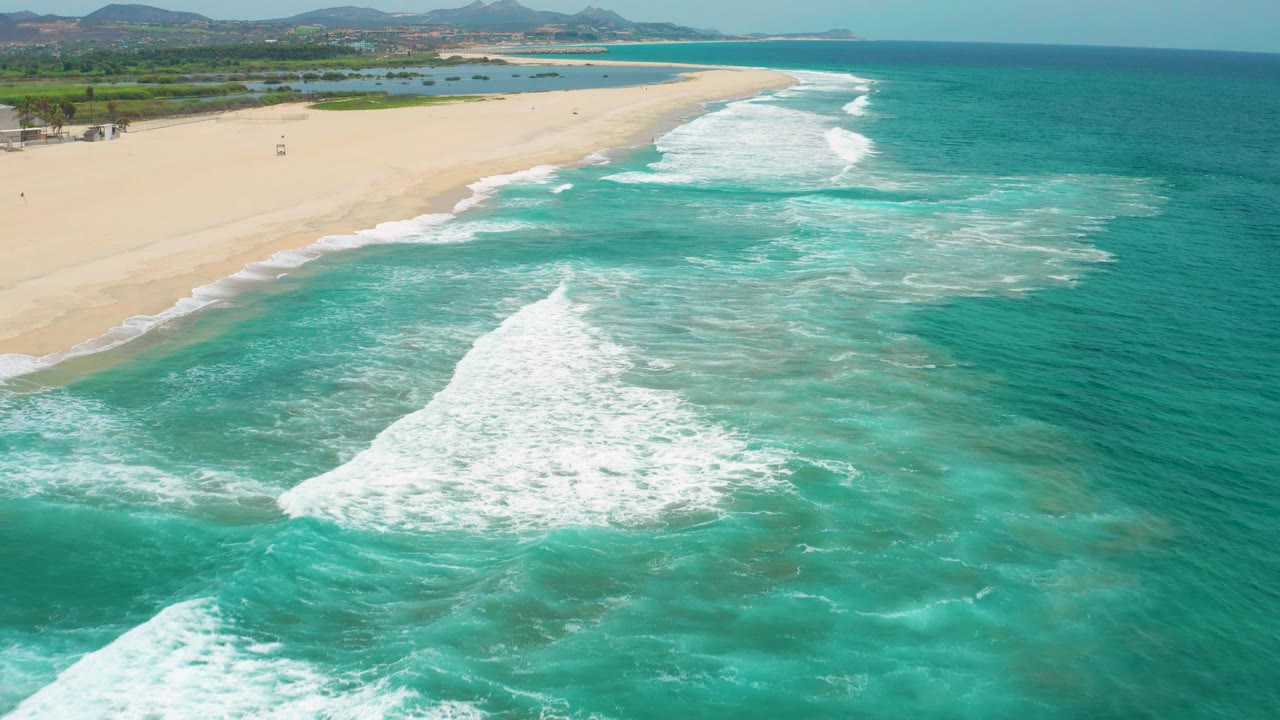 Aerial view in San Jose Del Cabo, Mexico with a large patch of white surf spreading over shallow turquoise water, the sandy beach along the left edge, hills in the distance, and no visible people.