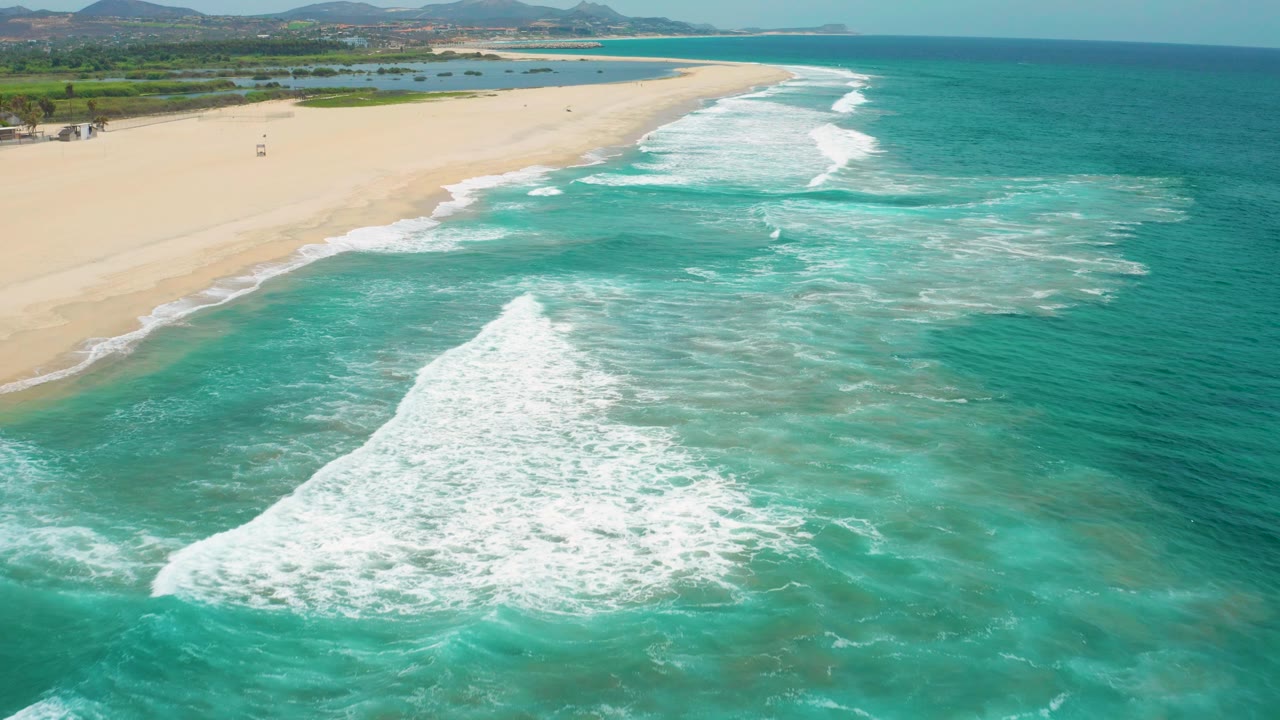 San Jose Del Cabo, Mexico coastline seen from above, with open turquoise water across the foreground, a wide foamy wave crest near center, empty sand to the left, and no visible people.