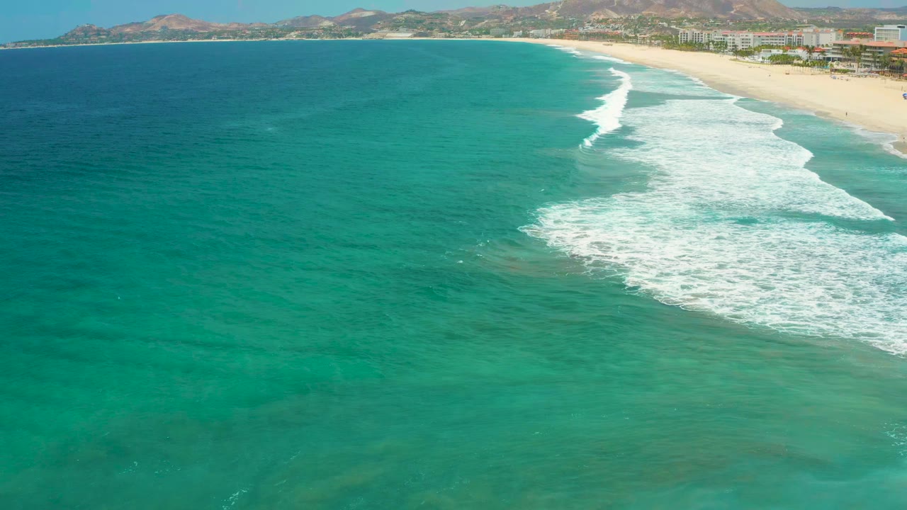 Aerial view over San Jose Del Cabo turquoise water as a white wave line curls toward the sandy beach, with distant hills and no visible people.