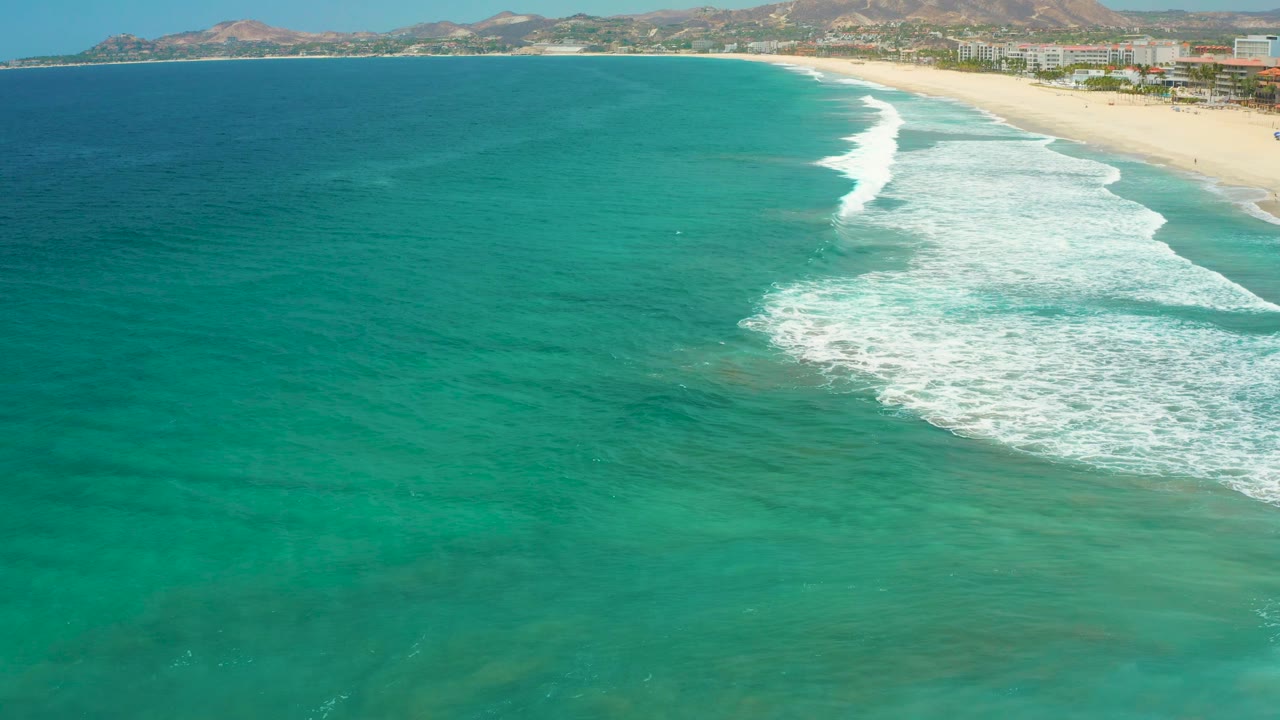 Aerial frame of San Jose Del Cabo clear green-blue water with foamy surf pushing shoreward, a pale beach at right, hills beyond the coast, and no visible people.